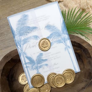 May include: Wedding invitation with blue palm tree design and gold wax seals with leaf imprints. The invitation includes text with wedding details. The seals and invitation are in a wooden bowl.