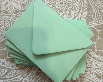 May include: A stack of mint green envelopes. The envelopes are triangular and have a smooth texture. The envelopes are stacked on top of each other, with the top envelope slightly open. The background is a patterned fabric.
