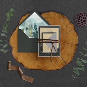 Puede incluir: Un conjunto de invitaciones de boda con una escena de monta&ntilde;a de acuarela en el frente. Las invitaciones est&aacute;n atadas con cordel y colocadas sobre una rodaja de madera.