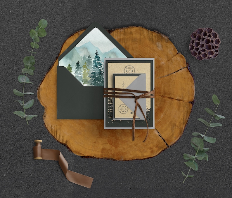 Puede incluir: Un conjunto de invitaciones de boda con una escena de monta&ntilde;a de acuarela en el frente. Las invitaciones est&aacute;n atadas con cordel y colocadas sobre una rodaja de madera.