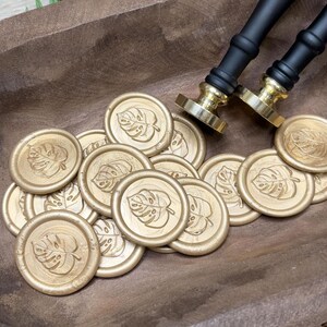 May include: Gold wax seals with a monstera leaf design are scattered on a wooden surface. Two black and gold wax stampers are in the background. The seals are round and have a detailed leaf imprint.