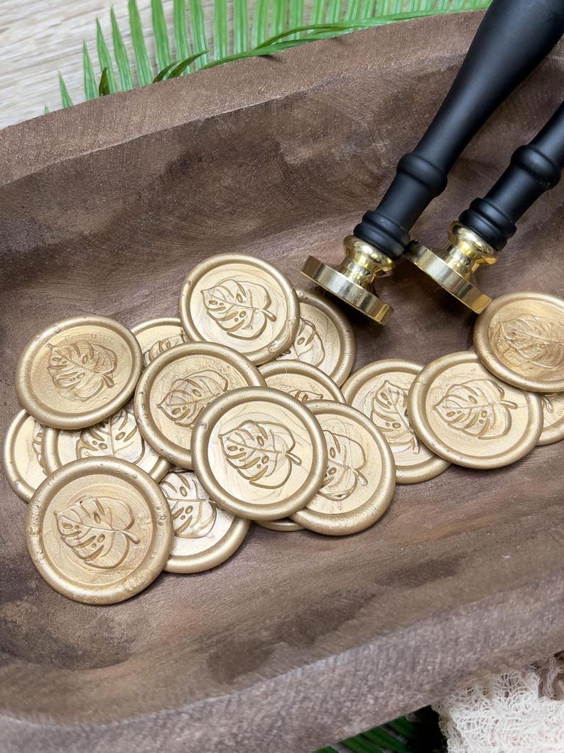 May include: Gold wax seals with a monstera leaf design are scattered on a wooden surface. Two black and gold wax stampers are in the background. The seals are round and have a detailed leaf imprint.