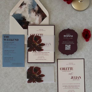 Burgundy, blush and dusty blue wedding stationery arranged in a layered flat lay, featuring a floral wedding invitation, scalloped RSVP card and coordinating details card with rich romantic colors.