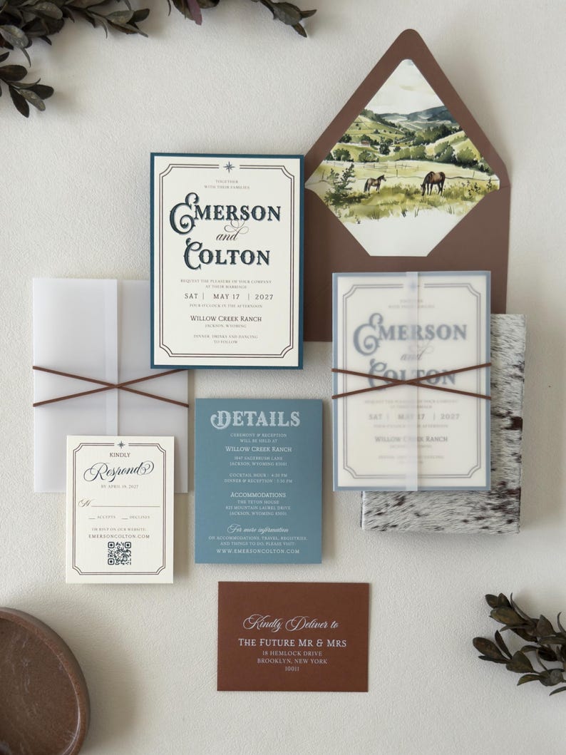 Western-inspired Emerson wedding invitation suite featuring an ivory invitation with vintage typography, dusty blue details card, square RSVP card, warm brown envelope and scenic ranch landscape liner.