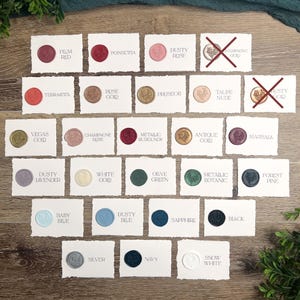 May include: A collection of wax seal samples in various colors, including Plum Red, Poinsettia, and Dusty Rose. Each seal is affixed to a small, white card with the color name printed below. The image showcases a variety of colors and finishes for wedding invitations or stationery.