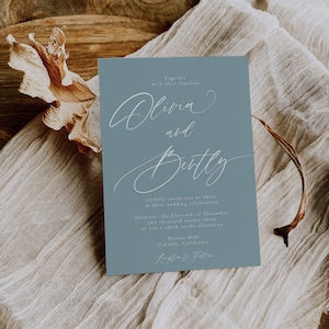 May include: A dusty blue wedding invitation with elegant white script. The invitation reads "Olivia and Betty" and includes event details. The card is set on a neutral linen cloth and wooden bowl, creating a rustic aesthetic.