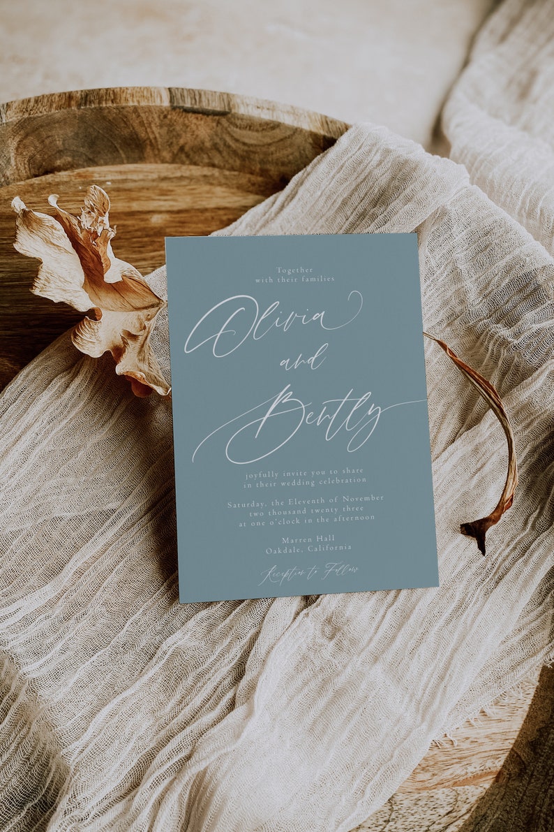 May include: A dusty blue wedding invitation with elegant white script. The invitation reads "Olivia and Betty" and includes event details. The card is set on a neutral linen cloth and wooden bowl, creating a rustic aesthetic.