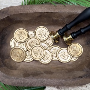 May include: Gold wax seals with a leaf design are scattered in a wooden bowl. Two black and gold wax seal stamps are positioned on the right. The seals and stamps are for crafting and stationery projects.