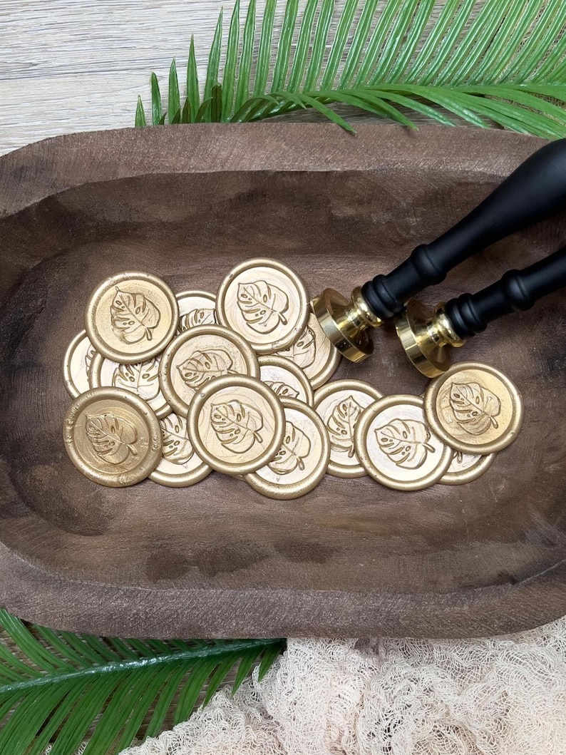 May include: Gold wax seals with a leaf design are scattered in a wooden bowl. Two black and gold wax seal stamps are positioned on the right. The seals and stamps are for crafting and stationery projects.