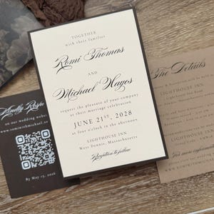 May include: A wedding invitation suite featuring a cream-colored main card with black serif and script text announcing the marriage of Romi Thomas and Michael Hayes on June 21st, 2028, at the Lighthouse Inn, accompanied by a dark brown RSVP card with a QR code.