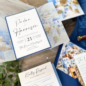May include: A wedding invitation with a blue and white color scheme. The invitation features a watercolor illustration of a Greek village with white buildings and blue accents. The text on the invitation reads "Padma and Harrison" and "October 21, 2026, 4:00 PM, The Barnal Countryside, Aurora, Oregon".