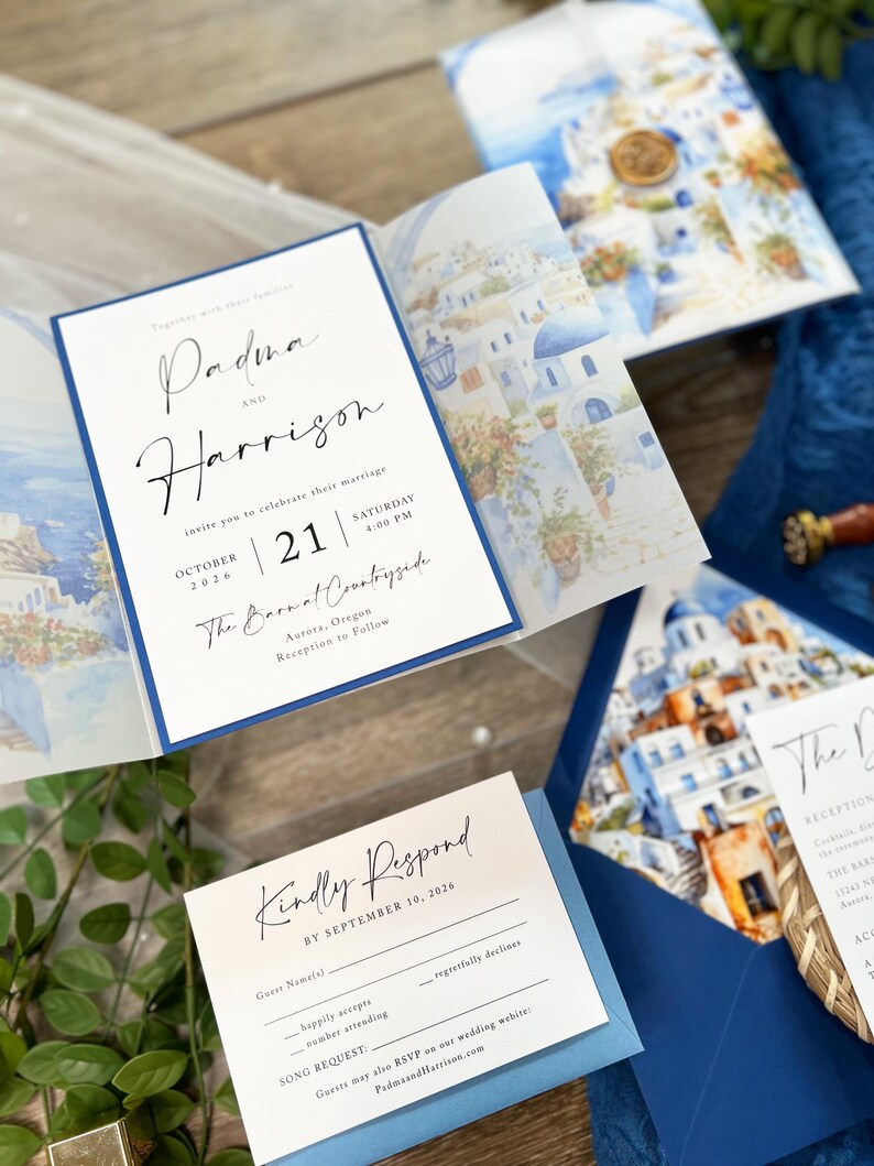 May include: A wedding invitation with a blue and white color scheme. The invitation features a watercolor illustration of a Greek village with white buildings and blue accents. The text on the invitation reads "Padma and Harrison" and "October 21, 2026, 4:00 PM, The Barnal Countryside, Aurora, Oregon".