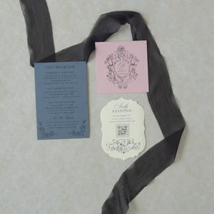 Odette wedding invitation accessories featuring a Marina blue details card, ivory botanical wedding invitation, scalloped RSVP website card, and Dusk pink decorative tag arranged with sheer black ribbon on a textured background.