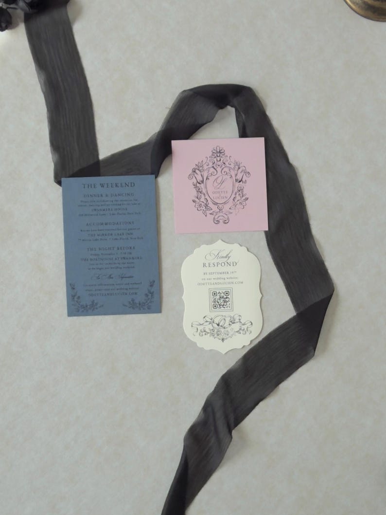 Odette wedding invitation accessories featuring a Marina blue details card, ivory botanical wedding invitation, scalloped RSVP website card, and Dusk pink decorative tag arranged with sheer black ribbon on a textured background.