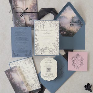 Odette enchanted swan lake wedding invitation suite featuring a Cloud Ivory invitation with Marina blue backing mat, illustrated misty woodland vellum jacket tied with black ribbon, Marina details card, scalloped RSVP, Dusk pink tag, and blue envelope with a romantic swan lake liner.