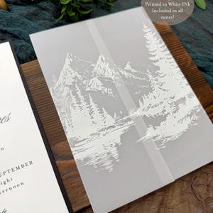 May include: A vellum jacket with a white mountain and forest design, printed in white ink. The jacket is part of a wedding invitation suite. The text on the jacket reads "VELLUM JACKET Printed in White Ink Included in all suites!"