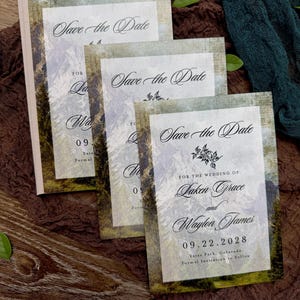 May include: A stack of wedding "Save the Date" cards with a mountain landscape design. The cards feature elegant script and the names Laken Grace and Waylon James, with the date 09.22.2028. The cards have a rustic, vintage aesthetic.