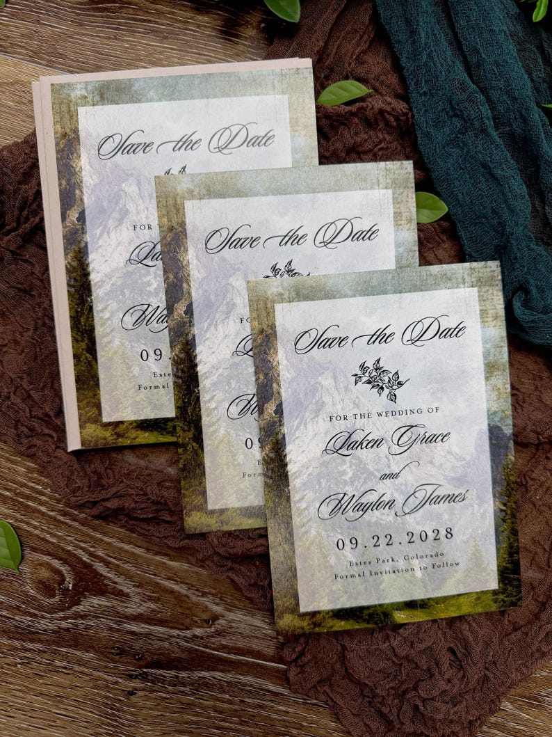 May include: A stack of wedding "Save the Date" cards with a mountain landscape design. The cards feature elegant script and the names Laken Grace and Waylon James, with the date 09.22.2028. The cards have a rustic, vintage aesthetic.