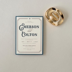 Ivory Emerson wedding invitation displayed with a warm brown envelope opened to reveal a scenic green ranch landscape liner and coordinating translucent stationery pieces.