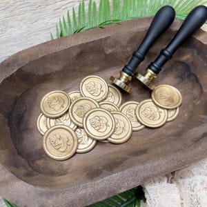 May include: Gold wax seals with a leaf design are scattered in a wooden bowl, alongside two black-handled wax stampers. The seals and stampers are arranged on a wooden surface with green palm leaves.
