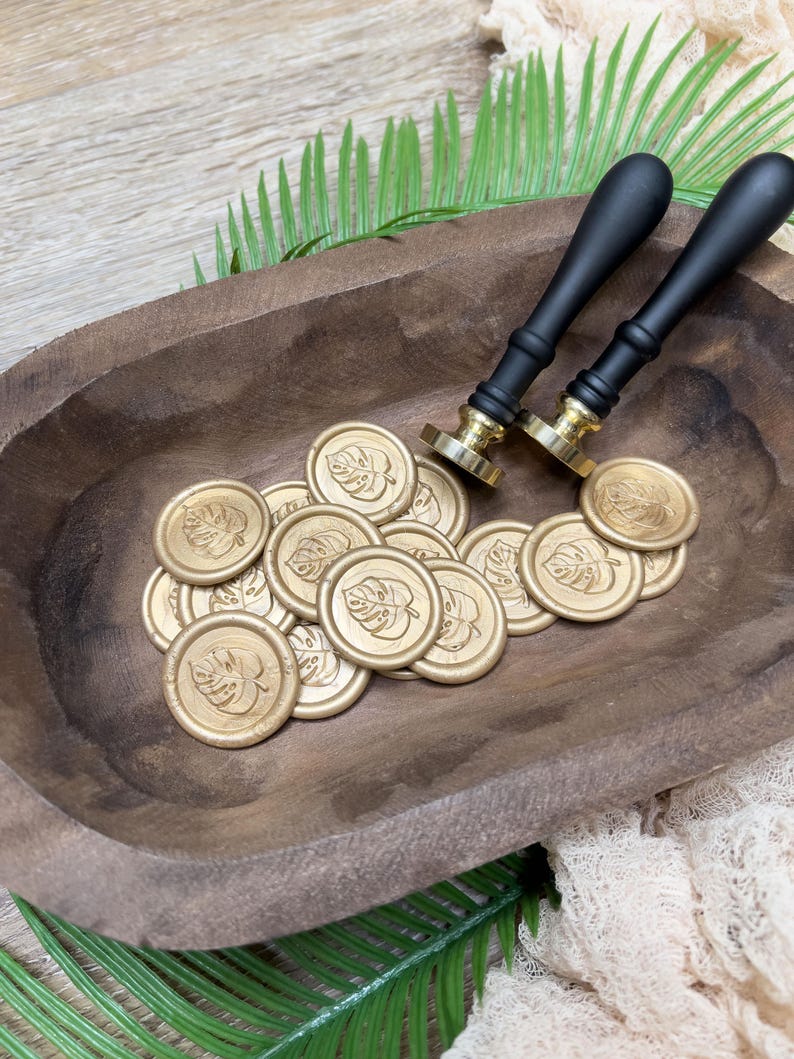 May include: Gold wax seals with a leaf design are scattered in a wooden bowl, alongside two black-handled wax stampers. The seals and stampers are arranged on a wooden surface with green palm leaves.