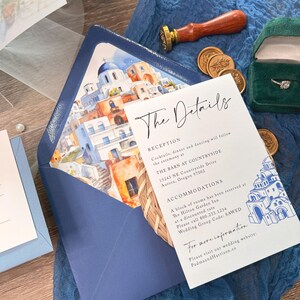 May include: A white wedding invitation card with blue and white watercolor illustrations of a Greek village. The card includes the text "The Details", "Reception", "Cocktails, dinner and dancing will follow the ceremony at", "THE BARN AT COUNTRYSIDE", "15243 NE Countryside Drive", "Aurora, Oregon 97002", "ACCOMMODATIONS", "A block of rooms has been reserved at", "The Hilton Garden Inn", "at a discounted rate", "Please call 800.233.1234", "Wedding Group Code: SAWED", "For more information", "Please visit our wedding website:", "PadmaandHarrison.co".