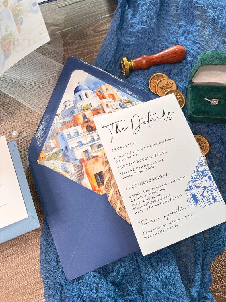 May include: A white wedding invitation card with blue and white watercolor illustrations of a Greek village. The card includes the text "The Details", "Reception", "Cocktails, dinner and dancing will follow the ceremony at", "THE BARN AT COUNTRYSIDE", "15243 NE Countryside Drive", "Aurora, Oregon 97002", "ACCOMMODATIONS", "A block of rooms has been reserved at", "The Hilton Garden Inn", "at a discounted rate", "Please call 800.233.1234", "Wedding Group Code: SAWED", "For more information", "Please visit our wedding website:", "PadmaandHarrison.co".
