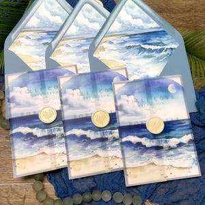 May include: Wedding invitation suite featuring watercolor artwork of a beach scene with ocean waves, sand, and sky. The invitations are sealed with gold wax seals and are presented with matching blue envelopes.