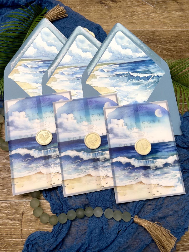May include: Wedding invitation suite featuring watercolor artwork of a beach scene with ocean waves, sand, and sky. The invitations are sealed with gold wax seals and are presented with matching blue envelopes.