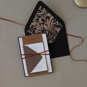Layered Whitmore wedding invitation wrapped with thin copper brown ribbon and displayed with a black envelope featuring an ornate botanical liner.