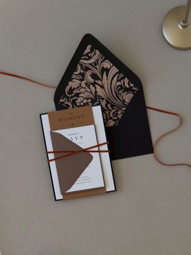 Layered Whitmore wedding invitation wrapped with thin copper brown ribbon and displayed with a black envelope featuring an ornate botanical liner.