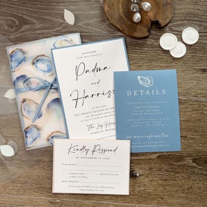May include: A wedding stationery set in blue and white. The set includes an invitation, details card, RSVP card, and a vellum wrap with oyster shell designs. The text includes the names Padma and Harrise.