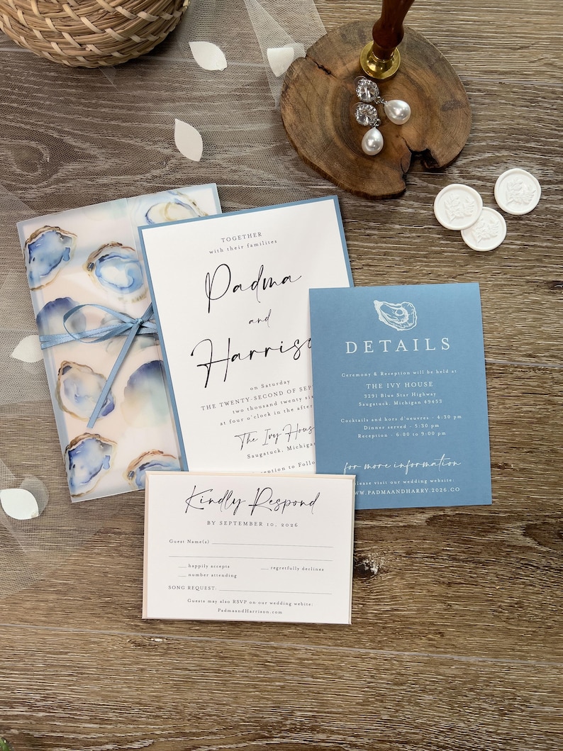 May include: A wedding stationery set in blue and white. The set includes an invitation, details card, RSVP card, and a vellum wrap with oyster shell designs. The text includes the names Padma and Harrise.