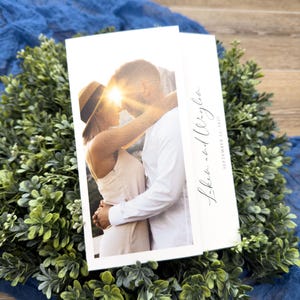 Könnte beinhalten: Eine Hochzeitseinladung mit einem Foto eines sich umarmenden Paares, die Sonne zwischen ihnen. Die weiße Einladung trägt die Namen "Laken and Wagner" und das Datum "September 22, 2023" in Schreibschrift. Umgeben von grünem Laub und blauem Stoff.