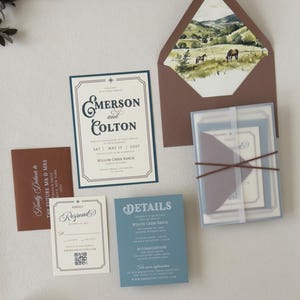Dusty blue and brown western wedding invitation suite featuring an ivory bordered invitation, blue details card, square RSVP card and brown envelope with scenic landscape liner.