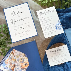 May include: Wedding invitation suite with a blue and white color scheme. The invitation features a white card with black calligraphy text that reads "Padma and Harrison" and "October 21, 2026, Saturday 4:00 PM, The Barn at Countryside, Aurora, Oregon". The details card features a blue and white illustration of a cityscape. The RSVP card features the text "Kindly Respond by September 10, 2026".