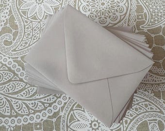 May include: A stack of light gray envelopes, with a triangular flap, are arranged on a patterned surface. The envelopes are made of a smooth paper material. The background features a beige fabric with a white floral design.