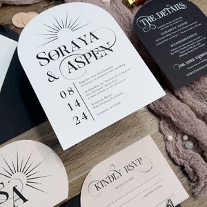 May include: Wedding invitation suite with a pink and black color scheme. The invitation features a sunburst design with the names "Soraya & Aspen" and the date "08.14.24". The RSVP card is pink with black text and a black envelope.