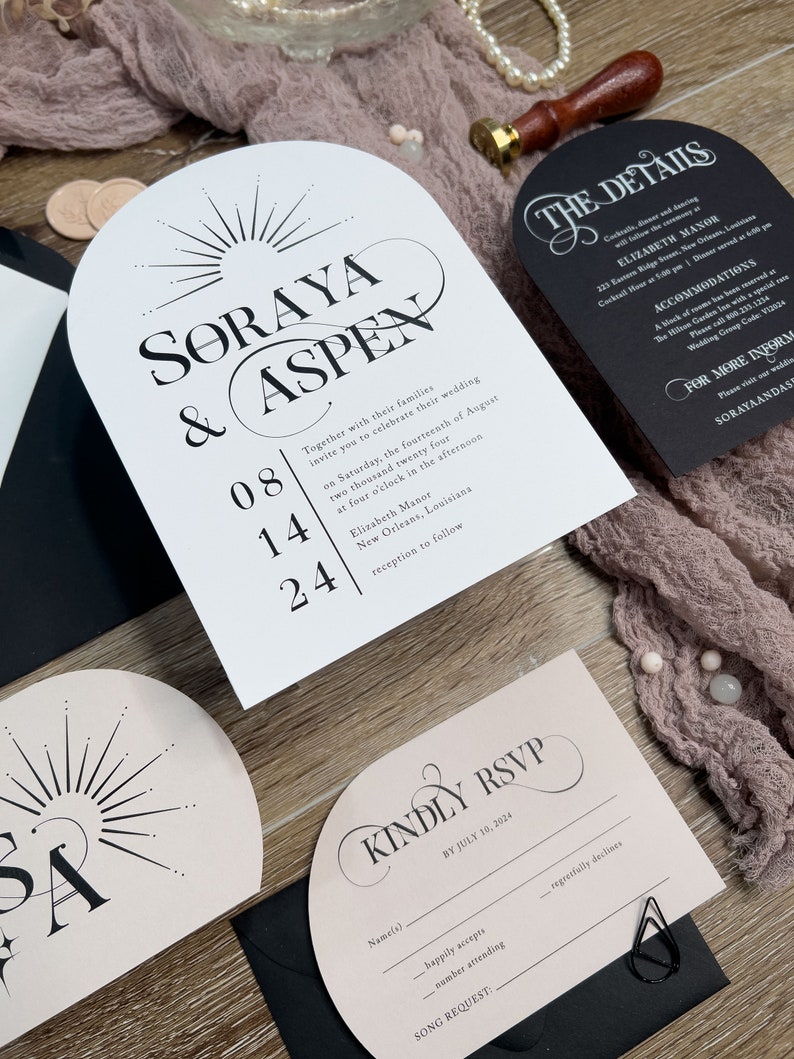 May include: Wedding invitation suite with a pink and black color scheme. The invitation features a sunburst design with the names "Soraya & Aspen" and the date "08.14.24". The RSVP card is pink with black text and a black envelope.