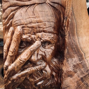 May include: A detailed wood carving of a man's face, with his hand covering his eyes. The carving is in shades of brown, with visible wood grain and intricate details. The artwork is a close-up shot, highlighting the texture and craftsmanship.