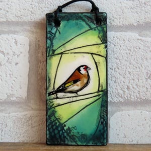 May include: Rectangular ceramic wall hanging with a bird illustration. The bird has a red head, brown body, and yellow details, perched on a branch against a green and yellow backdrop. A black cord is attached for hanging. The artwork is approximately 10 cm wide.