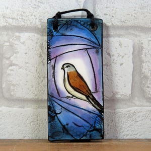 May include: Rectangular ceramic wall hanging with a painted bird design. The bird has a white chest, brown wings, and a black tail, set against a blue and purple background. A black cord is attached for hanging.