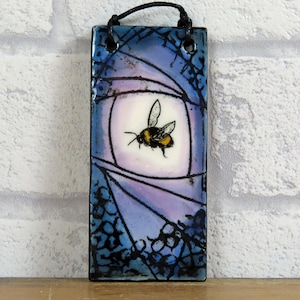 May include: Rectangular ceramic wall plaque with a bumblebee design. The artwork depicts a bumblebee with black and yellow markings against a soft, circular backdrop. The background is a blend of blue and purple hues, with a black cord for hanging.
