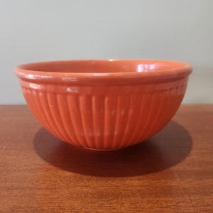 May include: An orange ceramic mixing bowl with vertical ribbed detailing. The bowl has a rounded shape and a smooth rim. The bowl is sitting on a wooden surface.