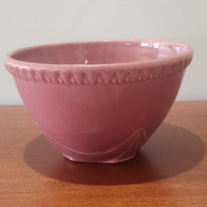 May include: A dusty rose-colored ceramic bowl with a decorative rim. The bowl has a rounded shape and a smooth, glossy finish. The rim features a repeating pattern of small, raised designs. This vintage bowl is suitable for kitchen use.