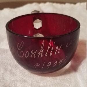 May include: A deep red glass cup with a clear glass handle. The cup has the inscription "Conklin" and "1909" etched into the side.
