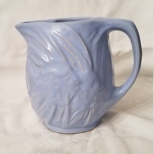 May include: A light blue ceramic pitcher with a textured design of leaves and flowers. The pitcher has a wide mouth and a handle.