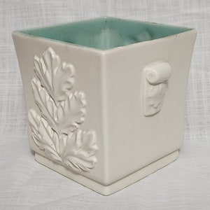 Puede incluir: Maceta de cerámica blanca con forma cuadrada y un glaseado verde claro en la parte superior. La maceta tiene un diseño de hoja en relieve en un lado y un asa en el otro.