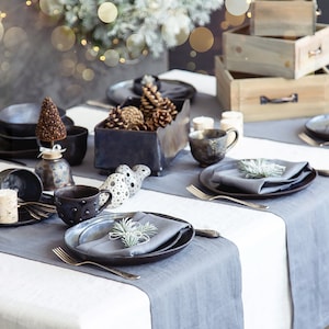 May include: A rustic table setting with a gray tablecloth and gray linen placemats. The table is set with black and gray ceramic dishes, a gray napkin, and a sprig of greenery. There are also small ceramic ornaments and a wooden box with pine cones.