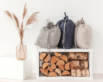 May include: Three drawstring bags in striped grey, solid navy blue, and solid beige fabric. The bags are positioned on a white shelf filled with stacked birch firewood logs. A clear glass vase containing dried pampas grass sits nearby.